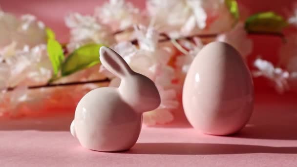 一只瓷制复活节兔子和蛋站在粉红的背景上 枝头上的苹果树枝条模糊不清 近景侧视 右边旋转着一圈 — 图库视频影像