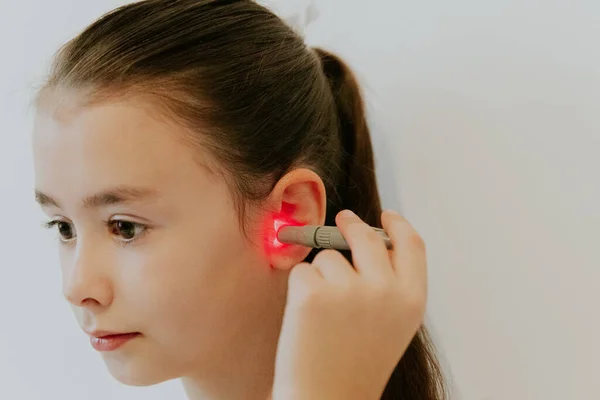 One beautiful little Caucasian brunette girl with her hair in a ponytail is treating her left ear with an infrared light device, sitting on the bed at home near a white wall, side view very close-up.