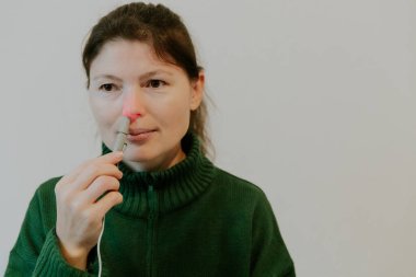 Yeşil kazaklı, beyaz saçlı, beyaz bir duvarın yanındaki bir yatakta oturan, kızıl ötesi ışıklı bir aletle sağ burun deliğini tedavi eden, beyaz saçlı, güzel bir esmer kız.