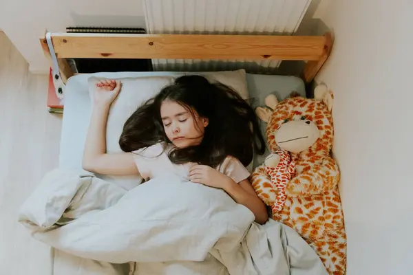 One beautiful little Caucasian brunette girl with flowing hair sleeps sweetly early in the morning in a wooden bed with a soft toy giraffe covered with a gray blanket near the radiator, close-up top
