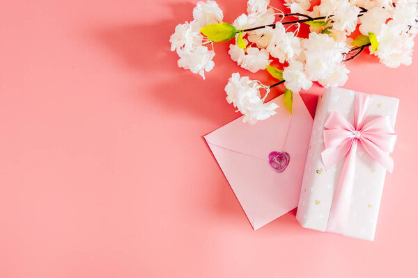 One beautiful gift box with a bow, a sealed envelope and branches of apple tree flowers lie on the right on a pink background with copy space on the left, flat lay close-up.