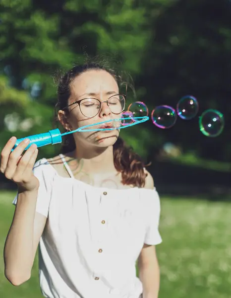 Portrait of one beautiful Caucasian teenage girl with closed eyes in glasses blowing round soap bubbles in a row, standing in a park on a playground, close-up side view. PARKS and RECREATION concept