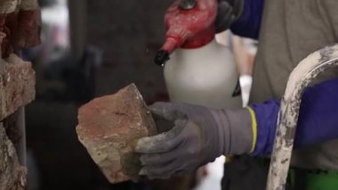 Sıradan mavi uzun kollu genç bir beyaz adam merdiven üzerinde duruyor ve bir tuğlayı sprey şişeden ıslatarak su ile ıslatıyor, tavanın altındaki bir kapıda duruyor.