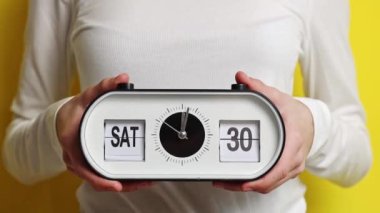 Elinde saat, tarih ve gün olan genç bir beyaz kız var: Cumartesi, 30 Mart Pazar ve 31 Mart 'a geçiyor, sarı bir arka planda asılı duruyor.
