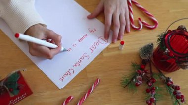 Beyaz kazaklı, tanınmayan beyaz bir genç kız, Noel Baba 'ya Noel süslemeleri olan ahşap bir masada oturan kırmızı keçeli kalemle bir mektup yazar.