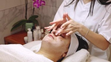 Kozmetik uzmanı güzel beyaz bir kız, bir güzellik salonundaki masaj masasında gözleri kapalı yatan yetişkin bir kadın müşteriye rahatlatıcı bir yüz masajı yapıyor.