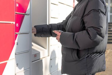 Siyah ceketli, tanınmayan genç beyaz bir adam elinde bir cep telefonu tutuyor ve diğeri de kutudan paketini almak için ekrandan bilgi giriyor.