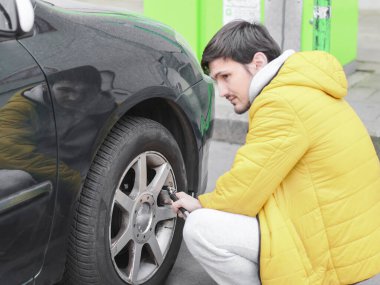 Sarı ceketli ve gri eşofmanlı genç beyaz adam, düşünceli bir şekilde çömeliyor, otoparkta hava kompresörü olan bir arabanın tekerleğine hava pompalıyor, seçici bir odak ile yan görüş ve