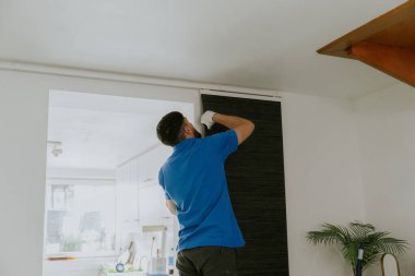 Mavi tişörtlü beyaz tenli genç bir adam yarım yamalak duruyor ve onları sökmek için koyu gri modern kağıt perdelere bakıyor..