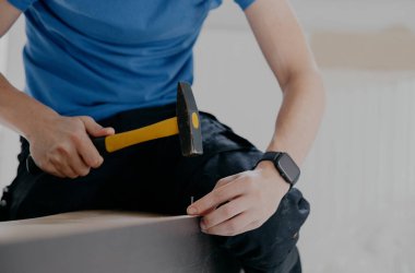 Mavi tişörtlü, siyah pantolonlu, tanınmayan beyaz bir genç, eski bir gardırobun arka duvarına sarı çekiçli bir çivi çakıyor..