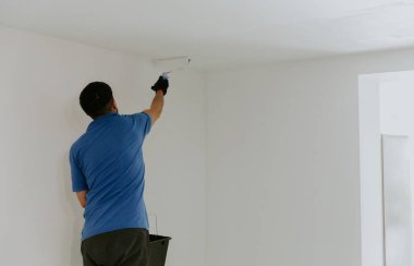 Mavi tişörtlü, kahverengi saçlı, genç bir beyaz adam arka tarafta duruyor ve tavanı beyaz boya rulosu ile yan tarafa boyuyor, seçici bir odak noktası var.