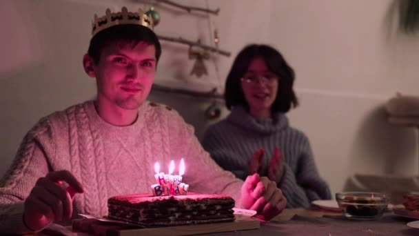 ある若いハンサムなハッピーな男が頭の上に黄金の紙の冠を持って 夕方のお祝いのテーブルに座っている間ケーキに幸せな誕生日のろうそくを吹き飛ばすために女の子のお祝いの歌を祝う — ストック動画