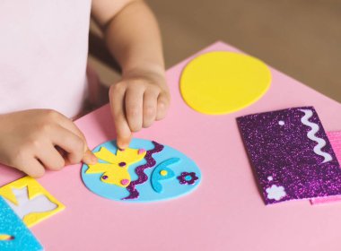 Kafkasyalı küçük bir kızın elleri sarı bir fiyonk yapıştırıyor parmakları mavi bir yumurta üzerinde bir şapka ve zigzag, pembe bir arka planda bir takım el sanatları ile bir çocuk masasında oturuyor