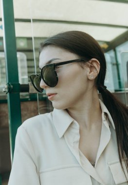 Güneşli bir bahar gününde halka açık bir parkta, kapalı bir kenar manzarada turist treninin açık vagonunda otururken, güneş gözlüklü ve şık giyinmiş güzel beyaz bir kızın portresi..