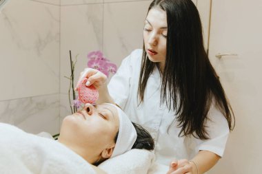 Genç, beyaz tenli, esmer bir kozmetik uzmanı tek eliyle, bir güzellik salonundaki masaj masasına gözlerini kapamış, yakından gören, pembe bir süngerle yetişkin bir müşterinin burnunu dikkatlice temizliyor..