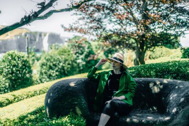 Hasır şapkalı, yeşil gömlekli, güzel beyaz bir kızın portresi. Tahta bir bankta, güneşli bir yaz gününde bulanık kayaların ve güzel doğanın arka planında oturuyor.