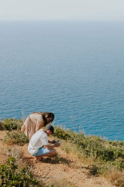Young Caucasian people, a guy and a girl, are watching a video they shot on a handheld phone on the top of a mountain against the backdrop of a blurred sea, close-up side view.