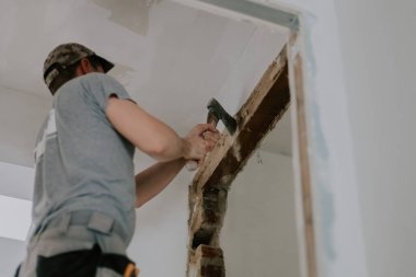 One young Caucasian man in a gray T-shirt and cap clears old putty from the top of a doorway with an ax while standing on a stepladder, close-up view from below with selective focus. Construction work