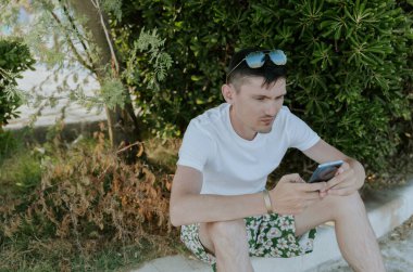 A portrait of one young Caucasian guy with sunglasses on his head sits on the curb near the bushes in a city park, enthusiastically communicates on a smartphone on a sunny summer day, close-up side