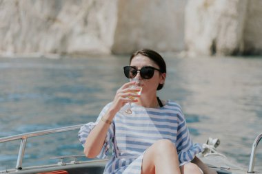 Portrait of one young Caucasian beautiful girl in sunglasses sitting in a boat and happily drinking from glasses of champagne against the background of blurred rocks while sailing on the sea on a