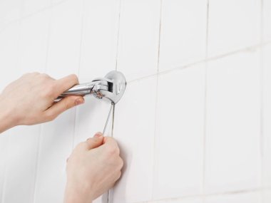 The hands of a young caucasian guy unscrew the screw with a small hex wrench to remove an old metal hanger with long tubes on a white tiled wall in the bathroom, close-up view from below. The concept