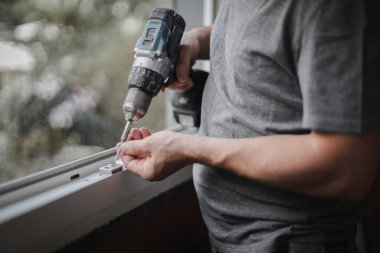 Young caucasian man holding drill and screw getting ready to install fittings on plastic window frame in renovation room, side view closeup with selective focus. The concept of home renovation