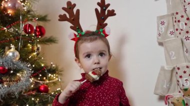 Kafasında geyik boynuzu çemberi olan iki yaşında güzel bir beyaz kız, duvardaki takvimin cebinden çıkartılan çikolatayı bir çubukla yer ve neşeyle midesini ovar.