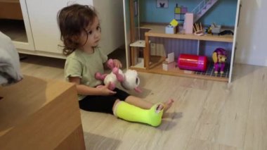 Sol bacağında açık yeşil bir alçı olan küçük, beyaz bir kız odada yerde oturuyor ve çocuk evinde plastik bir kedi oyuncağıyla oynuyor ve bacaklarını çaprazlıyor.