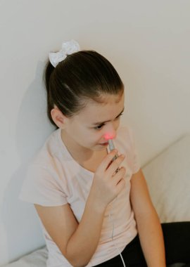 One beautiful Caucasian brunette girl with a serious emotion, collected hair and in a pink T-shirt treats the right nasal passage with a device with infrared light, sitting on the bed, cross-legged clipart
