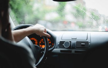 Arkadan oturan beyaz tenli genç adam yağmurlu bir günde, yakın çekim görüntüsüyle şehirde araba sürerken elini direksiyonda tutuyor. Araba sürme konsepti