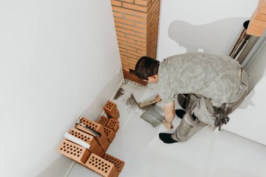Genç bir beyaz erkek müteahhit, bir süpürgeyle uzanan tuğlanın ortasında kurumuş çimento kalıntılarını süpürüyor ve odanın içinde bükülmüş vaziyette faraş gibi duruyor. Üst taraf görünümü kapalı. Temiz tuğla işi konsepti, ev yenilemesi..