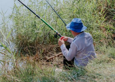 Kafasında mavi şapkası olan beyaz bir genç, güneşli bir yaz gününde, uzun bir çimenlikteki bir gölün kıyısında otururken oltaya bir solucan takar..