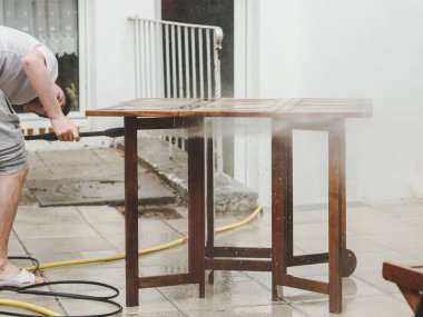 Gri eşofmanlı ve tişörtlü beyaz bir adam evinin arka bahçesindeki donmuş suyun basıncıyla ahşap bir bahçe masasını yıkıyor, aşağıdan manzara, yakın plan. Şey...