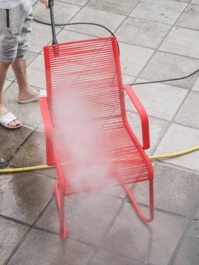 Gri eşofmanlı beyaz bir adam evinin arka bahçesindeki taş kiremitlerin üzerinde duran Karcher suyunun basıncıyla kırmızı bir tel sandalyeyi yıkıyor. Güneş yıkama kavramı