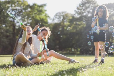 Three beautiful caucasian girls sisters are sitting on a flower lawn in a public park and blowing soap bubbles in the backlight with a blurred background, close-up side view. PARKS REC concept, happy