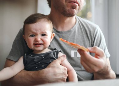 Babasının kollarında oturan beyaz bir kızın portresi babasının ona verdiği bir dilim ev yapımı pizzayı yemek istemediği için ağlayarak. Yuvarlak bir masada oturuyor.