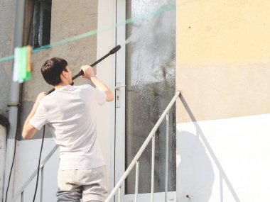 Gri spor giyen beyaz bir adam ve arkadan gelen bir tişört, evinin arka bahçesindeki metal bir merdivende dikilirken basınçlı bir karkasıyla pencereyi yıkıyor.