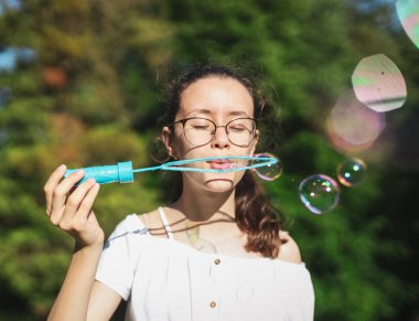 Güzel bir beyaz kızın portresi, gözlüklü mutlu genç bir kızın sabun köpüğü üflemesi, parkta dikilmesi, yakın çekimin alt tarafı. Parklar ve Rekreasyon Konsepti, Mutlu çocukluk