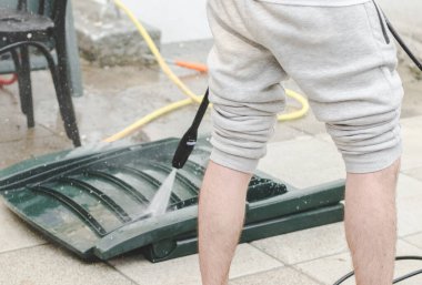 Gri eşofmanlı beyaz bir adam evinin arka bahçesinde katlanmış ve terasta uzanmış. Koyu yeşil güneş şezlongları. Karcher su jeti basıncının altında.
