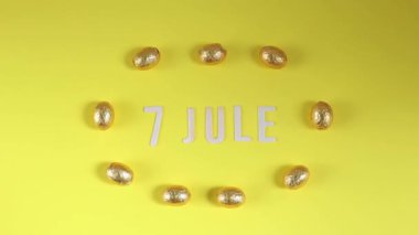 7 Temmuz ahşap harflerinin güzel manzarası. Ortasında güneşli sarı bir zemin üzerinde duran altın sarısı bir ambalajla sıralanmış dokuz çikolata yumurtası var. Yaklaş ve yakınlaş.