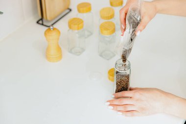 Tanınmayan genç bir beyaz kız plastik bir torbadan baharatlı siyah biberleri yeni bir cam şeffaf kavanoza döküyor. Yazın üst tarafında, mutfakta beyaz bir masada duruyor.