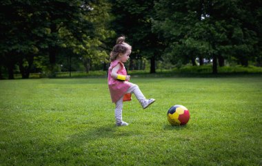 Bir yaz günü şehir parkında yeşil bir çimenlikte futbol topuyla oynayan beyaz elbiseli küçük bir kız çocuğu..