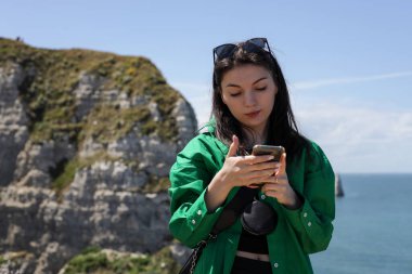 Yeşil tişörtlü, güzel, beyaz tenli bir kız Etretat 'ta güneşli bir yaz gününde bir dağın tepesinde dikilirken akıllı telefonunun ekranına bakar.