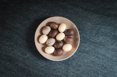 Beyaz, siyah ve sütlü çikolatalardan oluşan küçük çikolataların üzerinde güzel yumurtalar. Bir fincan tabağının içinde, koyu taştan bir arka planda, düz bir şekilde uzanıyorlar. Dünya Çikolata Günü konsepti.
