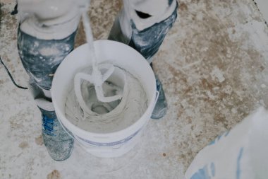 Genç, beyaz, beyaz, ıslak macunu beyaz bir kovayla karıştıran beyaz, beyaz, küçük bir kovada kirli bir zeminde duran, çuval içinde duran, üst tarafı kapalı, seçici bir odak noktası olan beyaz bir adam. Ev yenileme konsepti, inşaat işi.