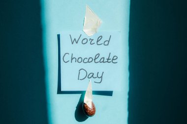 Dünya Çikolata Günü 'nün el yazısıyla küçük bir çikolatalı şeker yumurtası yapıştırılmış siyah bir kalemle koyu gölgeli mavi bir bantla yakın plan çekildiği mavi kareli bir not. Dünya Çikolata Günü konsepti.