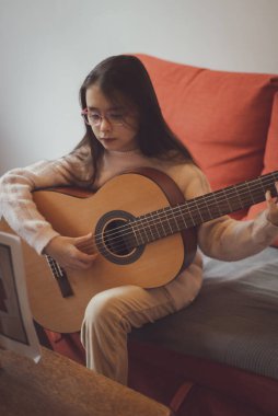 Kafkasyalı, güzel, uzun saçlı, gözlüklü esmer bir kız bir gitar tutuyor ve parmaklarıyla telleri koparıyor. Odadaki koltukta otururken notalar öğreniyor. Müzik eğitimi kavramı..