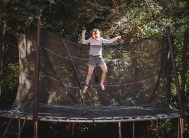 Şortlu güzel bir beyaz kız ve trambolinde kolları olan bir tişört, kolları havada bir evin bahçesinde, ağaçların arasında dikey bir şekilde dikilmiş, garip bir şekilde donmuş. Ev, ev sporları, mutlu bir çocukluk.