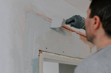 Gri üniformalı ve eldivenli genç bir beyaz esmer, inşaat ızgarasını spatula ile kaplıyor, kapı girişinde yarı açık duran bir merdiven, aşağıdan seçici bir odak ile yakın görüş. Ev kavramı