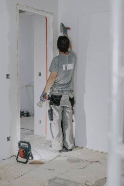 Gri üniformalı ve eldivenli yakışıklı beyaz bir adam, sırtından ayakta duruyor, akşamleyin duvarları küçük bir spatulayla kaplıyor ve bina feneri açık, yan görüş açısında. İnşaat konsepti.